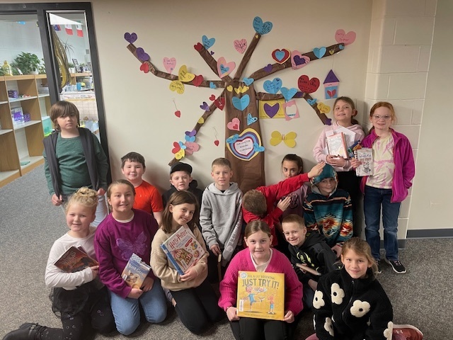 Elementary 3rd and 4th graders sitting in front of a Valentine's tree made out of hearts for leaves holding books
