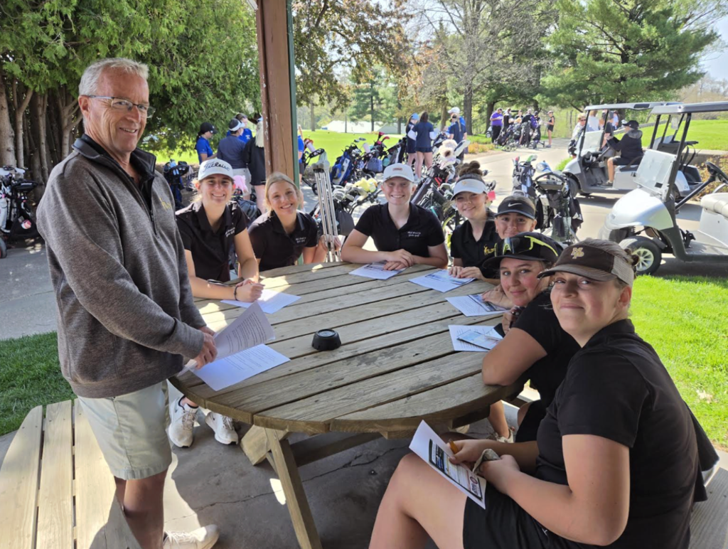 Girls golf team sitting at a table together with golf carts in the background