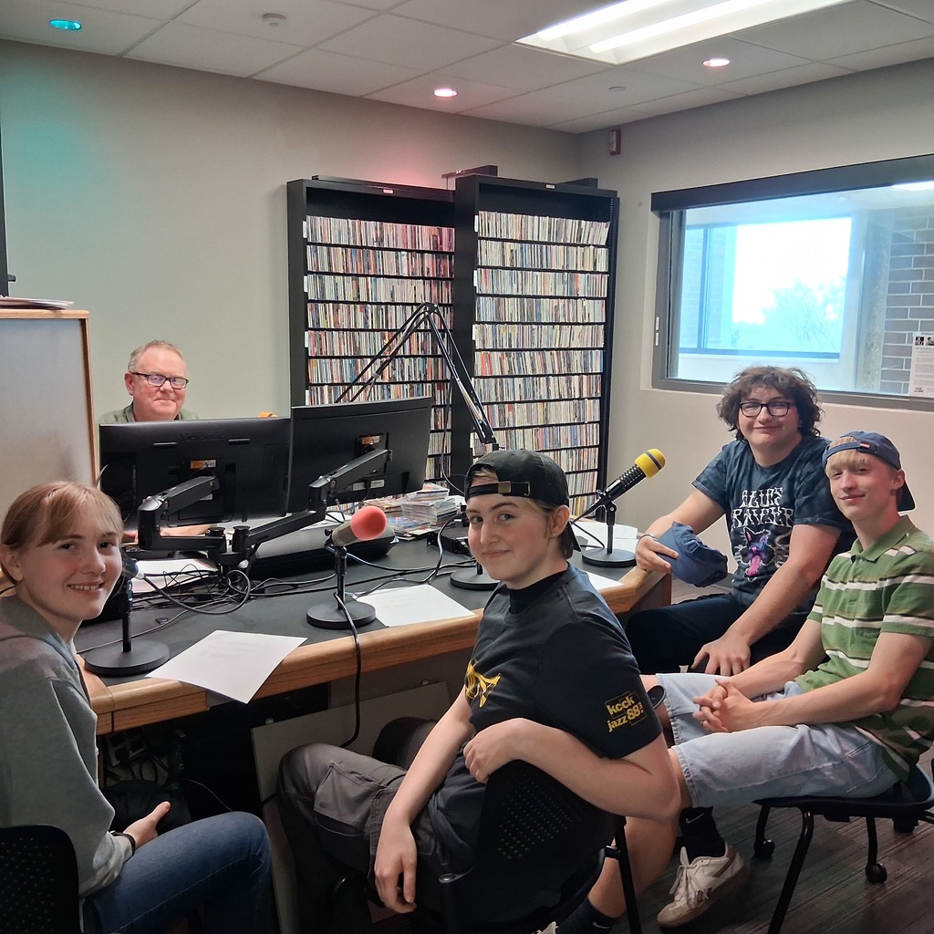 4 MPHS jazz band students sitting in the radio room with the DJ around a desk smiling with microphones in front of each one