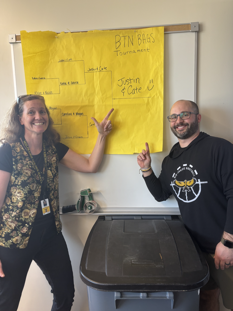 Two MP Teachers Cate Burke and Justin Barthelman pointing to a bracket competition drawn on a large piece of paper hanging on the wall showing that they made it to win the championship; Poster reads BTN BAGS Tournament with names in each player space and Cate and Justin written on the final champion line