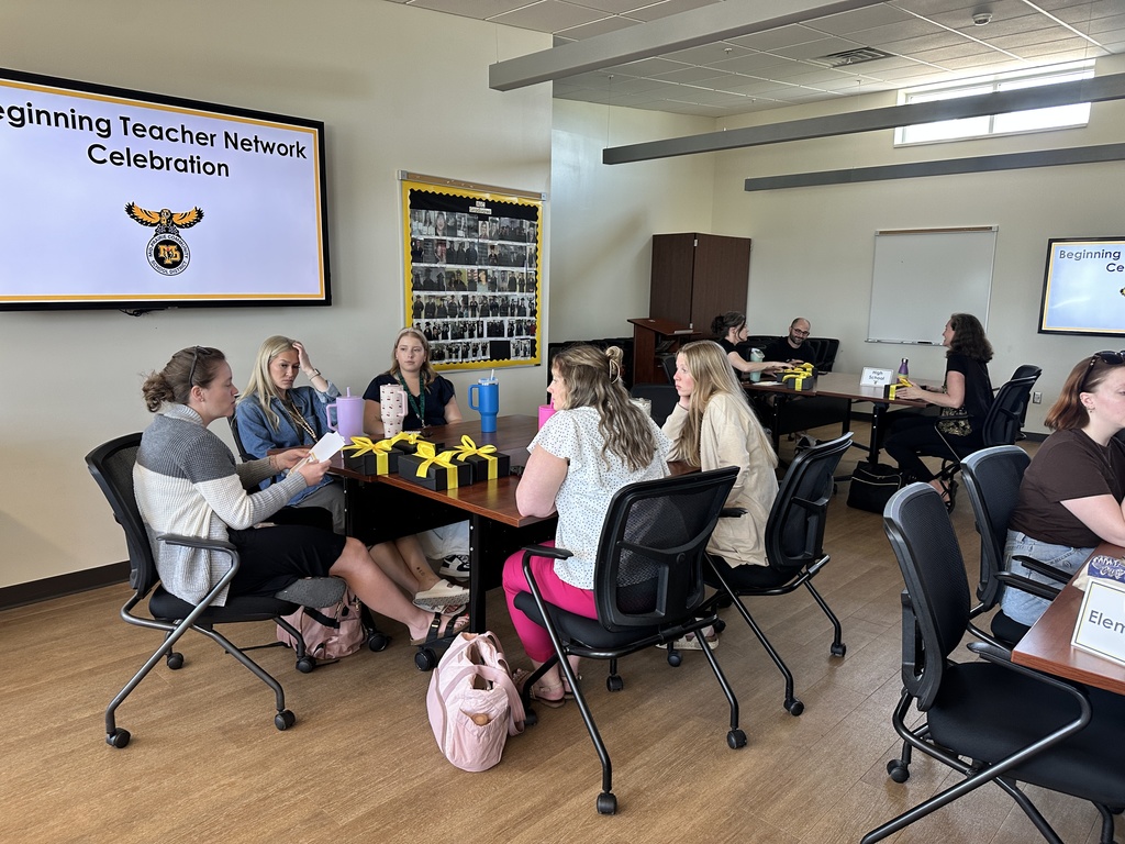 Groups of 5 teachers sitting at tables in the MP Central Office community room with gift boxes on the table in front of them; Text on the video screen reads Beginning Teachers Network Celebration