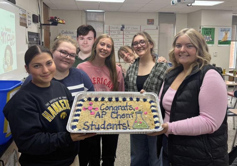 Group of high school students holding a cookie cake that says Congrats AP Chem Students with a picture of a science beaker with green frosting inside; Students are smiling, one student's sweatshirt reads The. University of Iowa Hawkeyes and another sweatshirt reads American