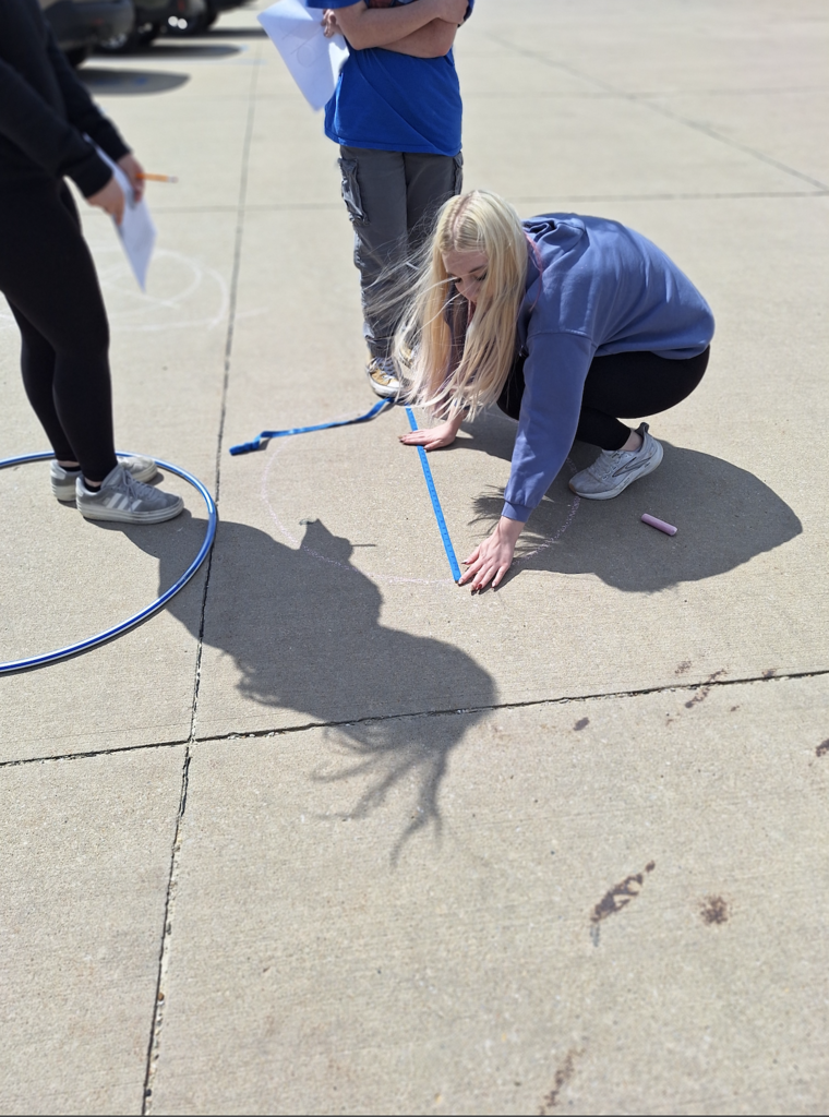 Female student using a measuring tape to measure a circle drawn with chalk on the parking lot pavement with two students standing next to her watching