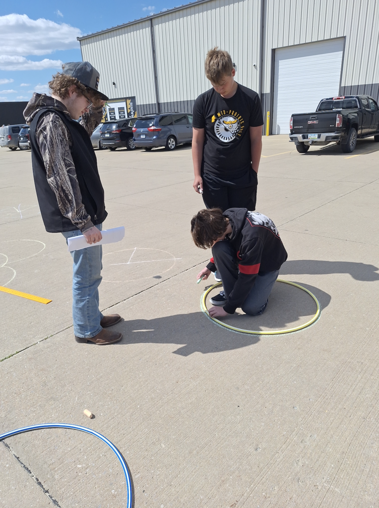 One male student tracing a circle with a hula hoop and chalk on the parking lot pavement with two male high school students standing next to him watching