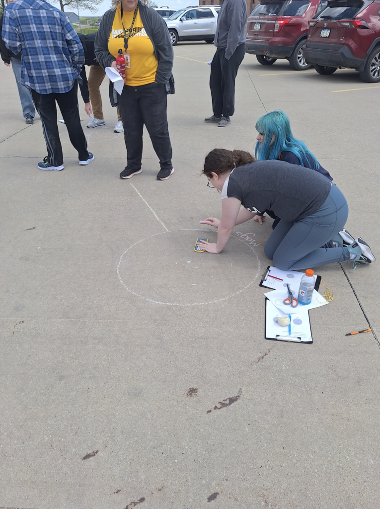 Male and female high school students drawing circles with chalk on the parking lot pavement with adults standing nearby watching