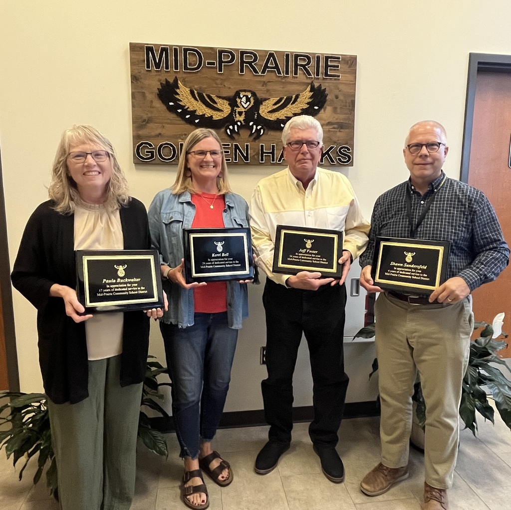 Paula Buckwalter, Kerri Bell, Jeff Foster, and Shawn Sandersfeld holding their retirement plaques in front of the sign that says Mid-Prairie Golden Hawks