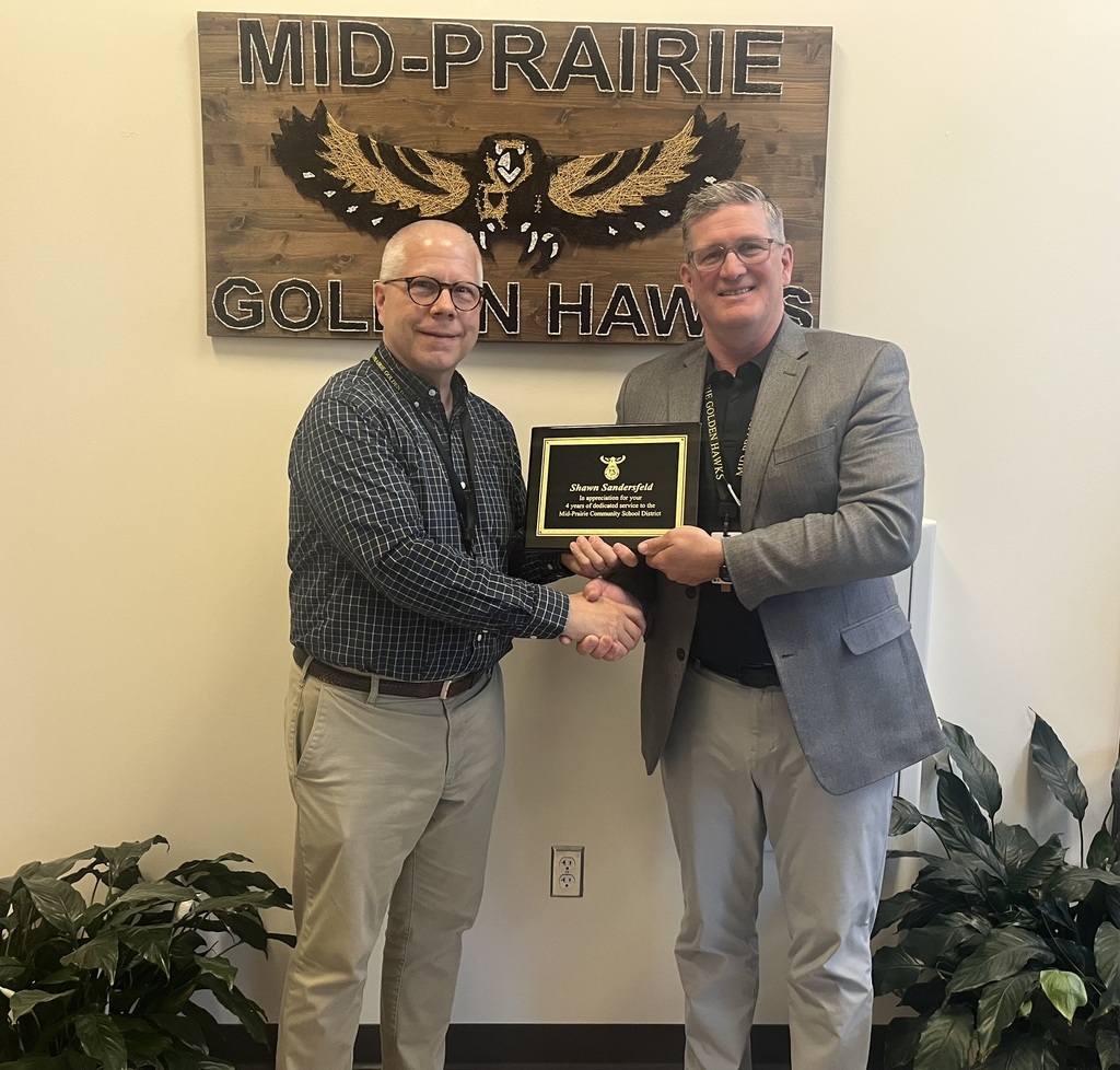 Shawn Sandersfeld and Brian Stone shaking hands holding the retirement plaque in front of the Mid-Prairie Golden Hawks sign