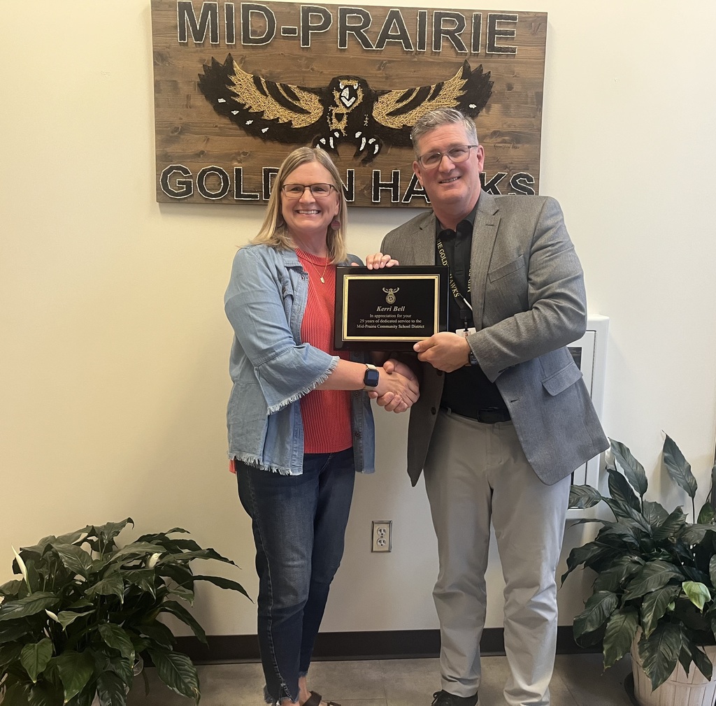 Kerri Bell and Brian Stone shaking hands holding the retirement plaque in front of the Mid-Prairie Golden Hawks sign