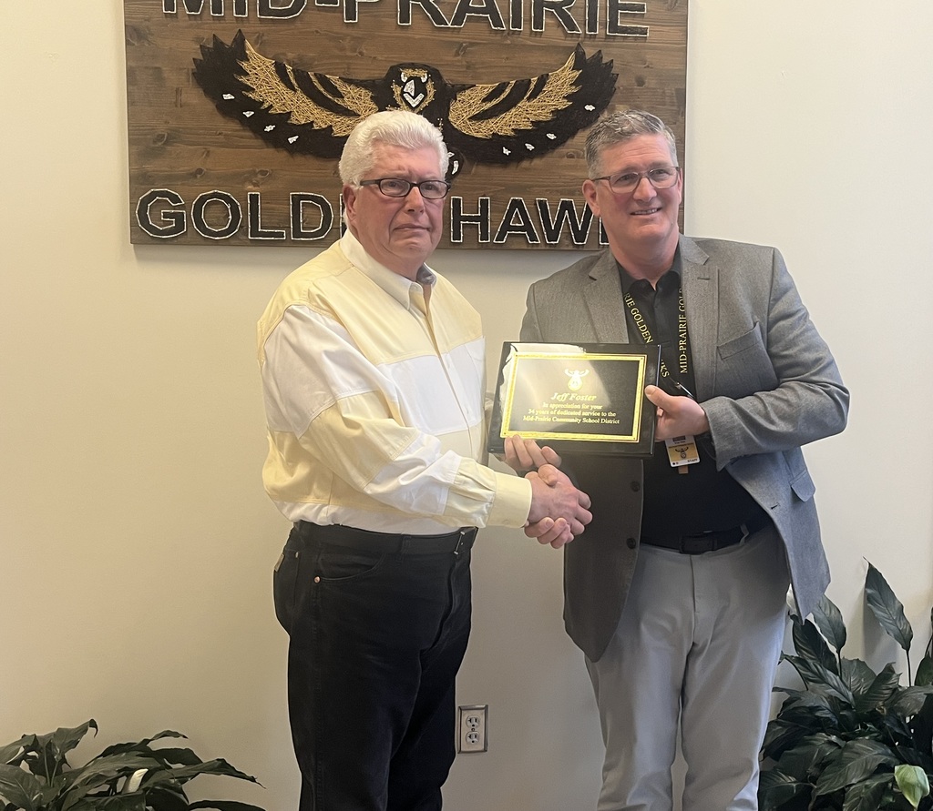 Jeff Foster and Brian Stone shaking hands holding the retirement plaque in front of the Mid-Prairie Golden Hawks sign