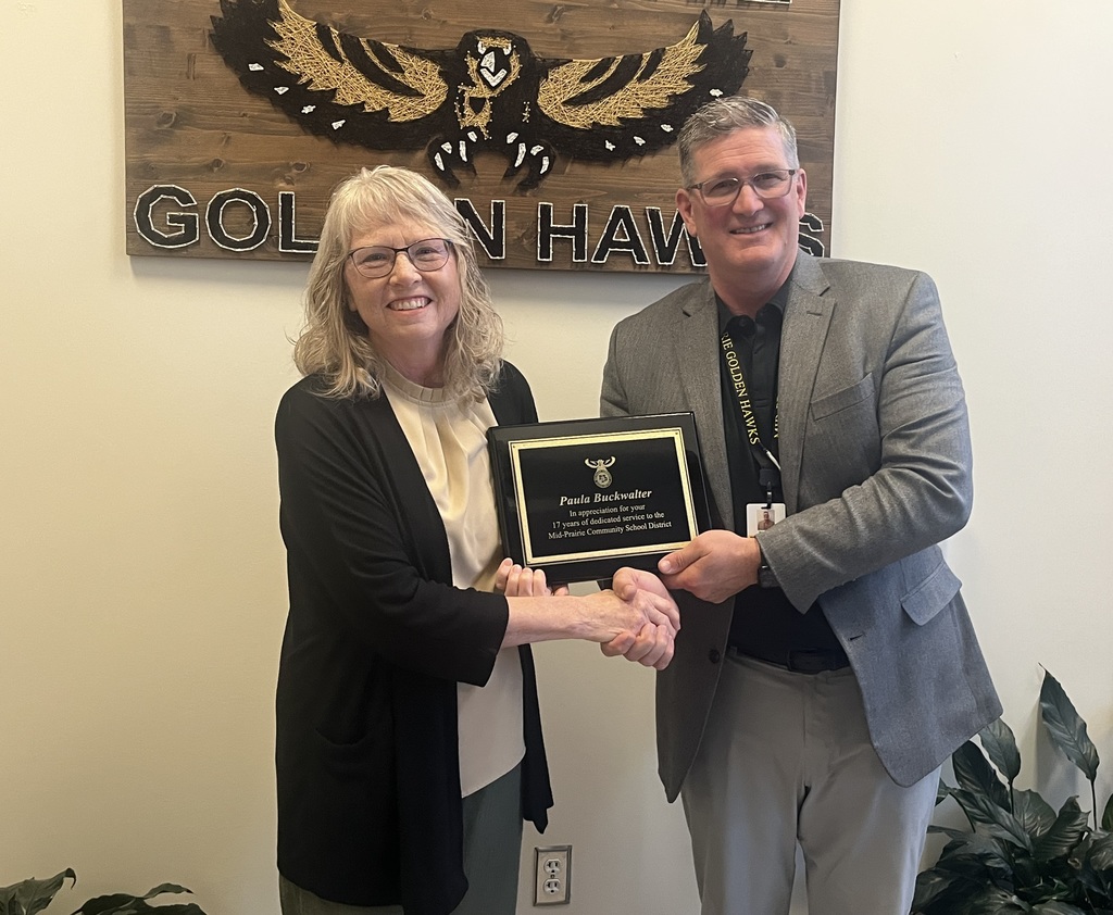 Paula Buckwalter and Brian Stone shaking hands holding the retirement plaque in front of the Mid-Prairie Golden Hawks sign