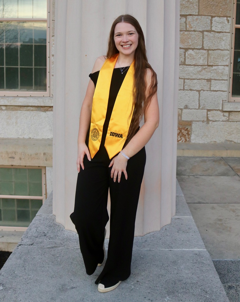 Claire Haag posing in black pants and shirt with gold Iowa graduation sash that says IOWA on it and has the UIowa emblem