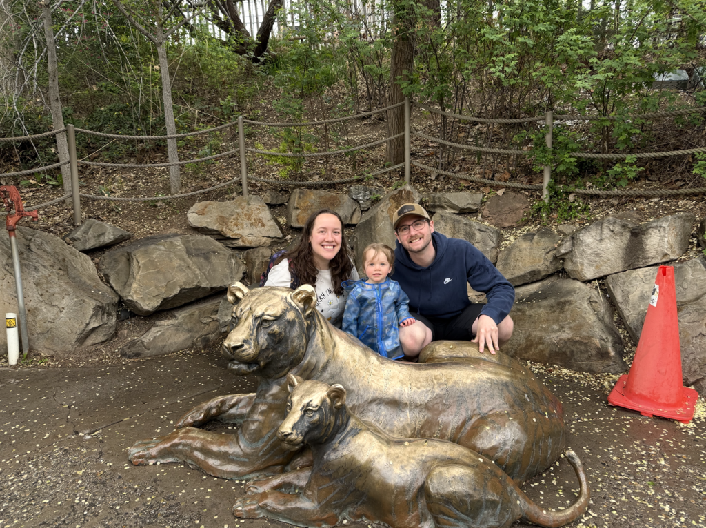Michaela Haskell with husband and daughter posing in front of two golden lions at a zoo