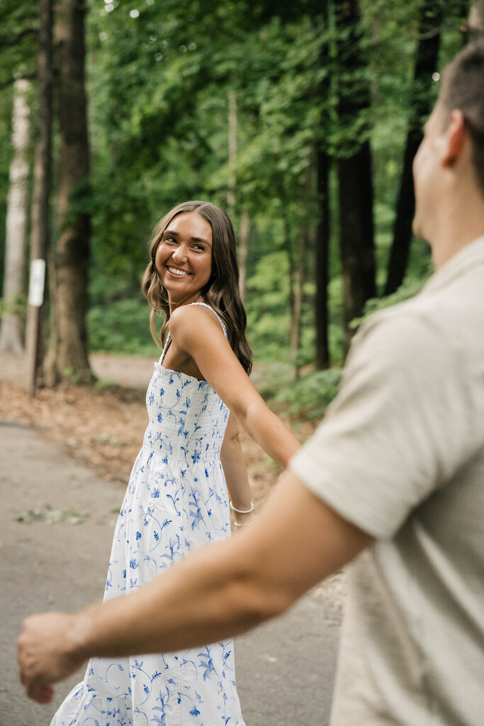 Kendall Burk with fiance walking through a forest path and she is wearing a white and blue flower dress looking back at him