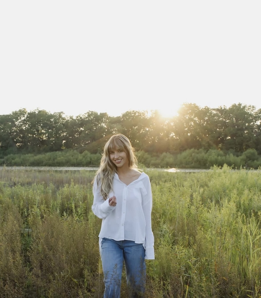 Madelyn Cunningham standing in a grassy field wearing a white long sleeved shirt and jeans smiling