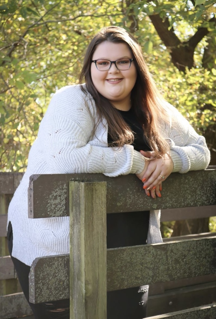 Hannah Collins standing on a wooden bridge with trees in the background wearing a white sweater and smiling
