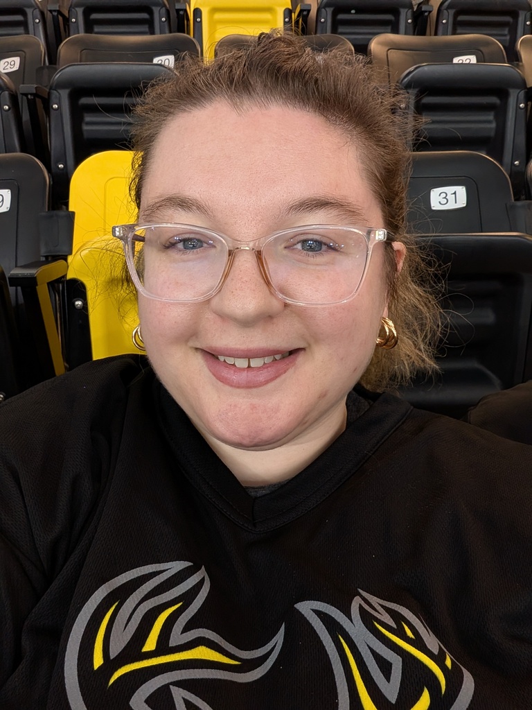 Aubrey DePew at a sporting event with black and yellow chairs behind her and wearing a black shirt and glasses smiling
