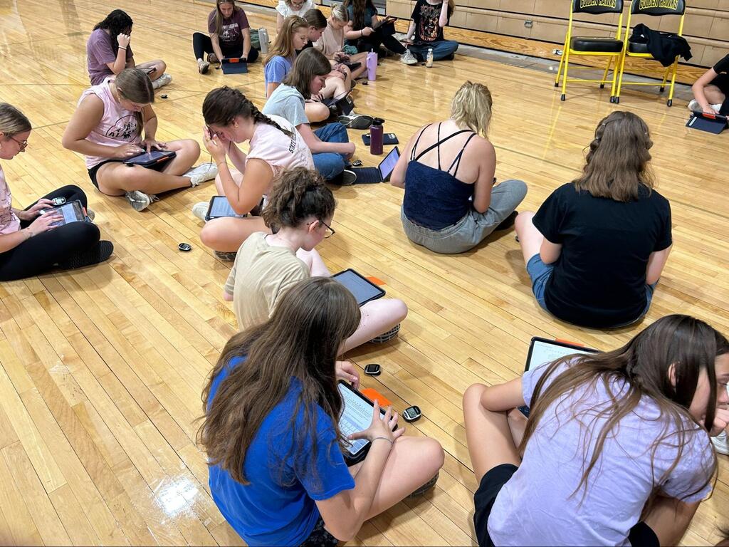 Students sitting on the gym floor cross-legged with ipads in their laps looking at them