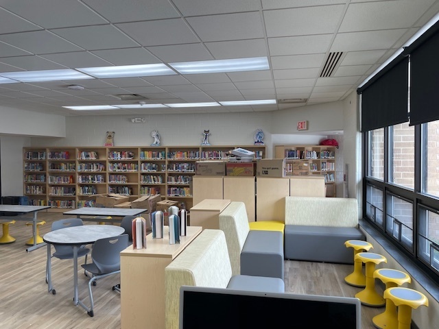Library room with shelves full of books lining the back wall, circular tables and rolling chairs, modern couches, and wobbly stools for students to sit on. All in coordinating colors of grey and gold.