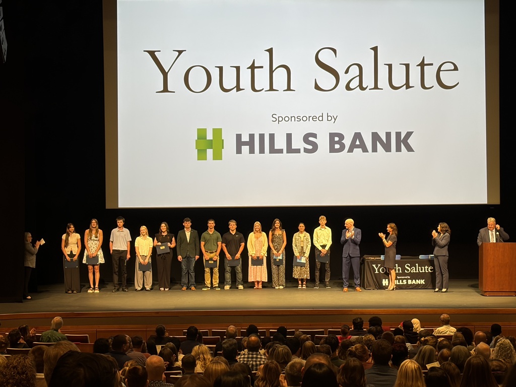 Mid-Prairie students on stage receiving their awards with a large screen behind them that reads Youth Salute Sponsored by Hills Bank: Maëlys Beachy, Morgan Bender, Eli Conrad, Gabbi Geno, Adelyn Huston, Caedyn Huston, Max Kipp, Tommy Miller, Taylor Pence, Brooklyn Schneider, Brooklyn Stutzman, Max Swartzendruber.