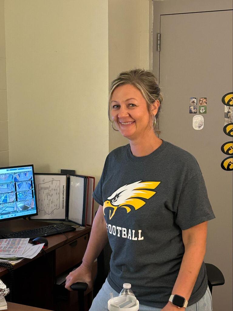 Melissa Rediger standing at her desk in front of a computer wearing a grey shirt that says MP Football