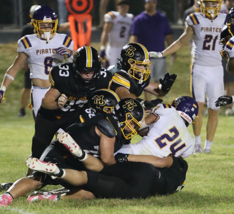 3 MP football players tackling a player in white jersey on the field