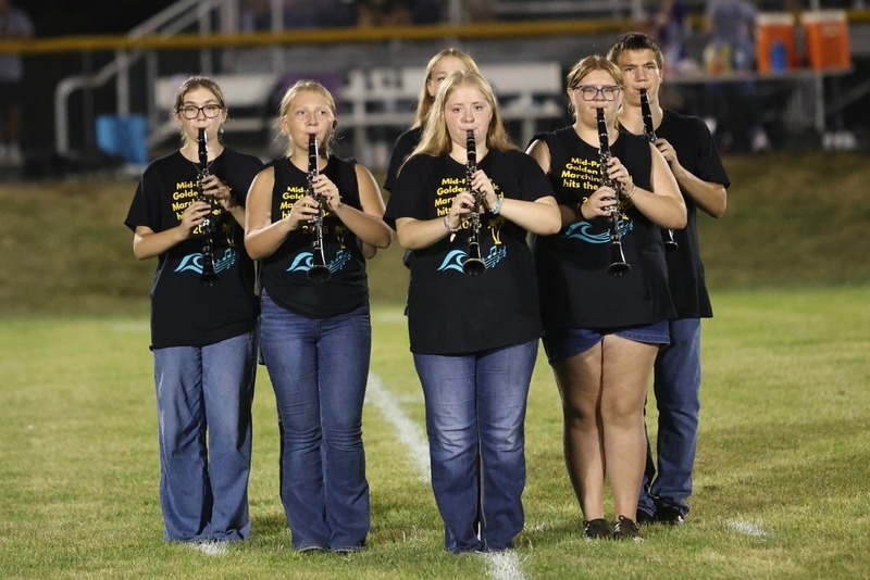 6 MP marching band students standing on the football field holding clarinets playing at half time; wearing black shirts with gold writing on it that says Mid-Prairie Golden Hawk Marching Band