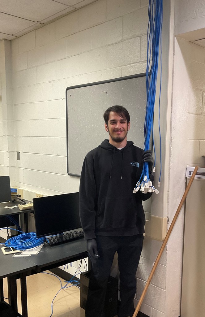 kyle he's kind of dressed in black he's standing in front of a monitor he has a little of wires and cables in his hand his smile on his face