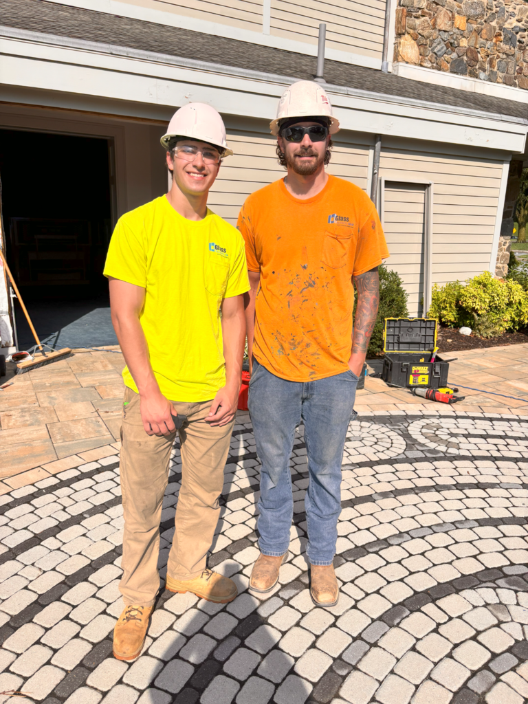 this picture of jason and ryan ryan's in a yellow shirt and a white hard hat Jason's in an orange shirt and a white hard hat but most safety glasses on they're standing on top of the pavers