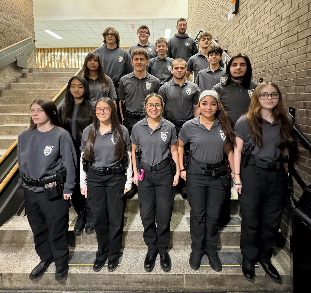 Take a photo of all of the morning session public safety students. They have black pants on and their uniform shirts standing on the stairs with their teacher.