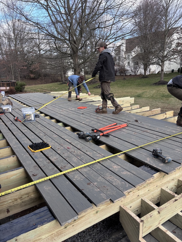 This is a picture of Aiden. He's on a work site. He's standing on some decking. He is wearing a black coat and a hat. He's outside. My hand looks like he's working with a pole. On a deck with some other guy stand.