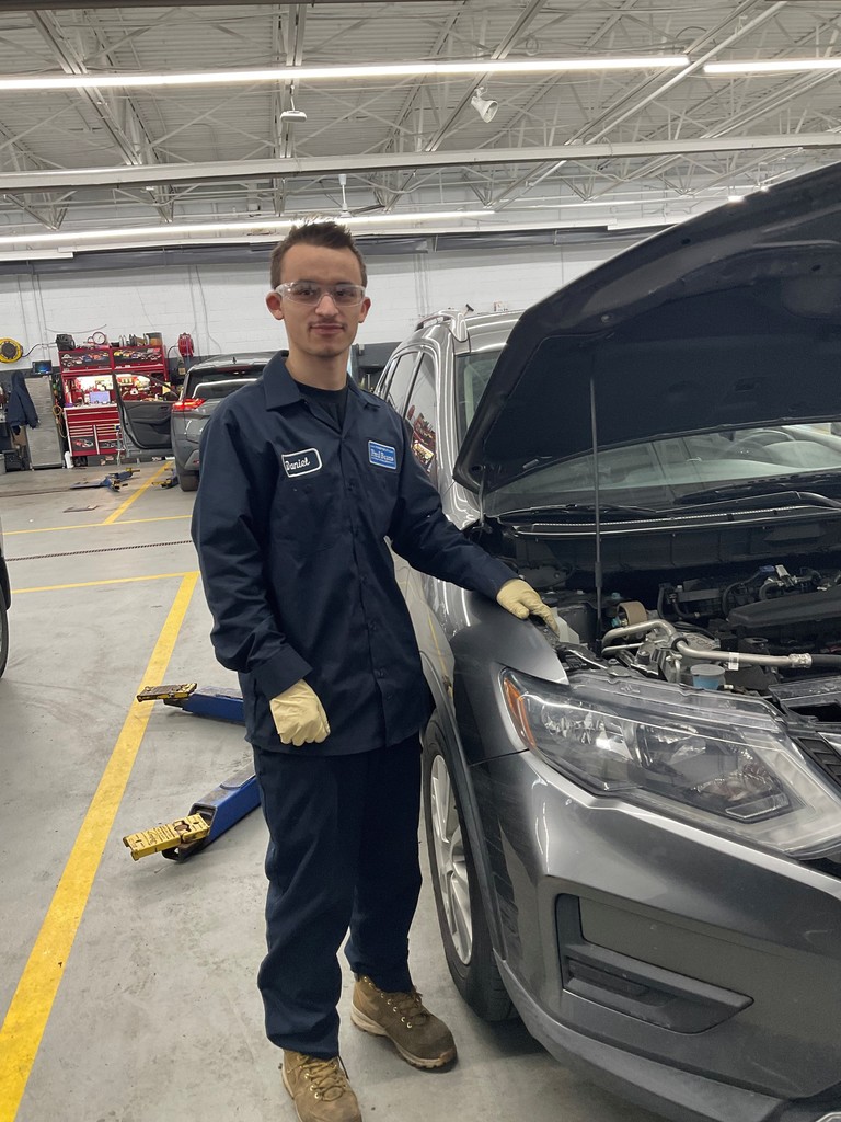 This is an image of Dan. He's in a automotive. Shop. He's got his Blues. on. He's working underneath the hood. He's got a smile on his face. He's got gloves on. He has safety glasses on