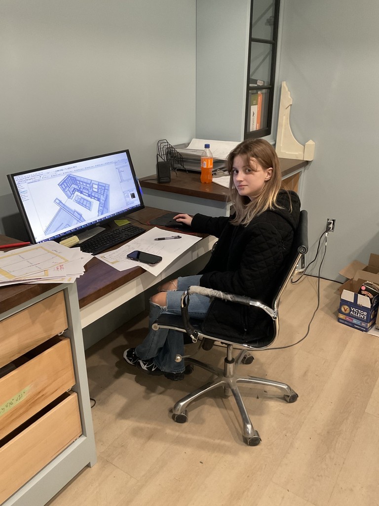 Picture of Michaela sitting at a desk with a black sweatshirt. She's working on a 3D image of a kitchen at her desk.