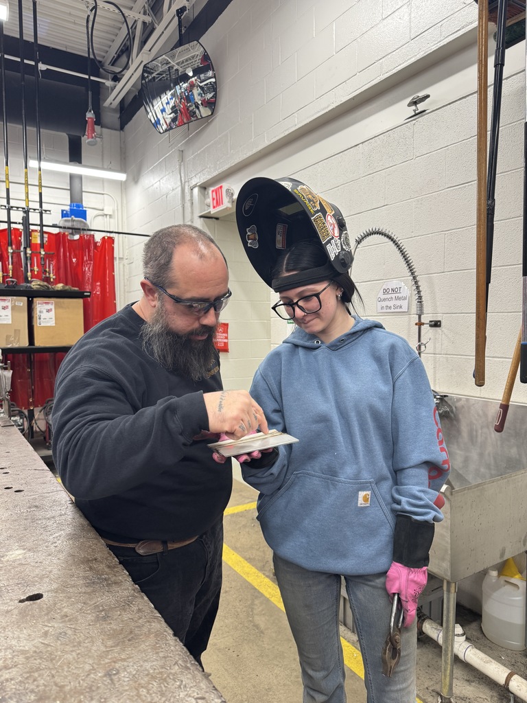 The teacher and the student, she's got a blue sweatshirt on it and a welding helmet flipped up. He is in a black sweatshirt. Safety glasses. They're looking at a piece of metal. She's got pink gloves and