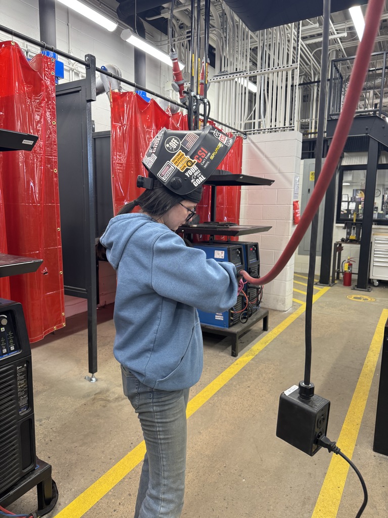 It's a female welding student. She's got a welding helmet flipped up blue sweatshirt and jeans on