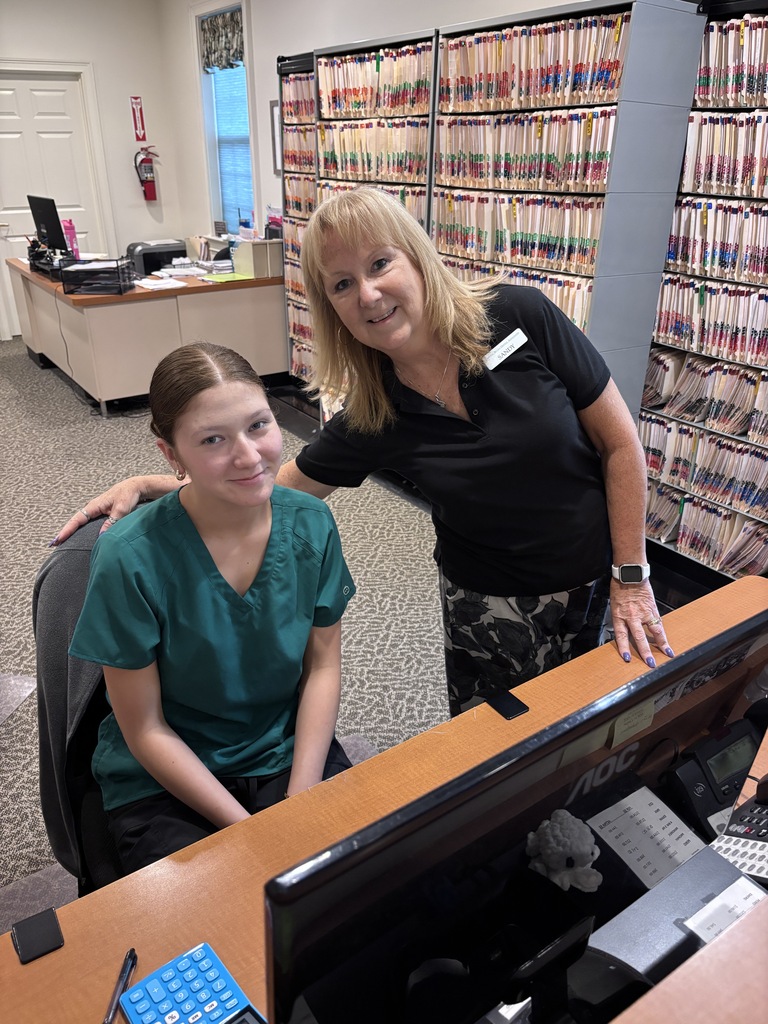 A picture of Abby she's sitting at the front desk at the dental Associates dental Associates. She's got scrubs on smile on her face, and one of her coworkers has her arm around her with a smile on her face.