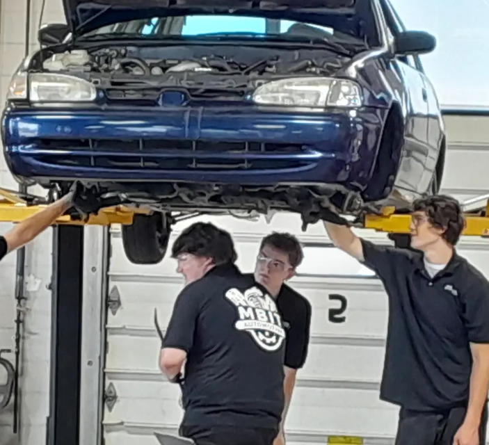 Multiple students in an automotive program with their uniforms on working underneath the car