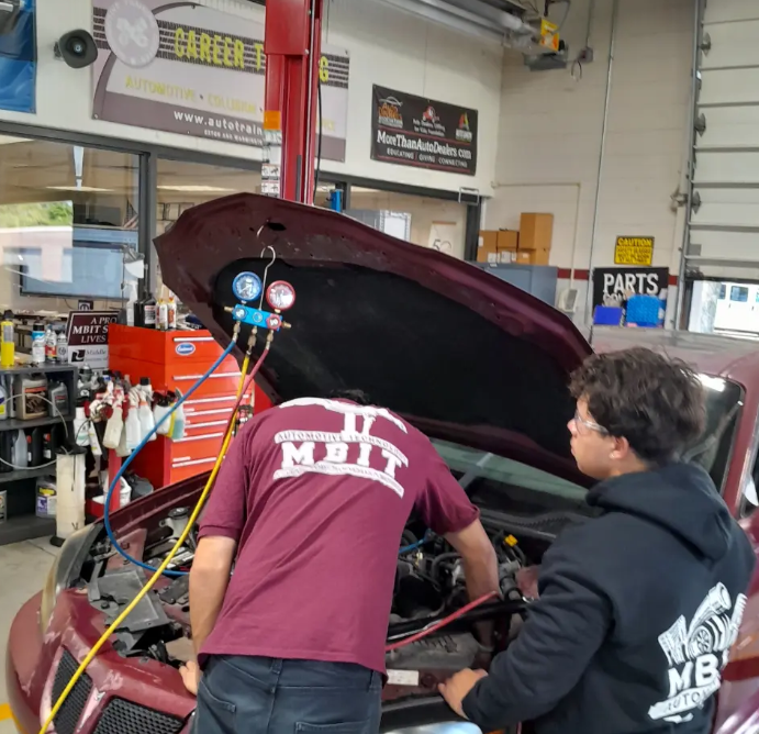 Two students working underneath the hood of a car in the automotive program