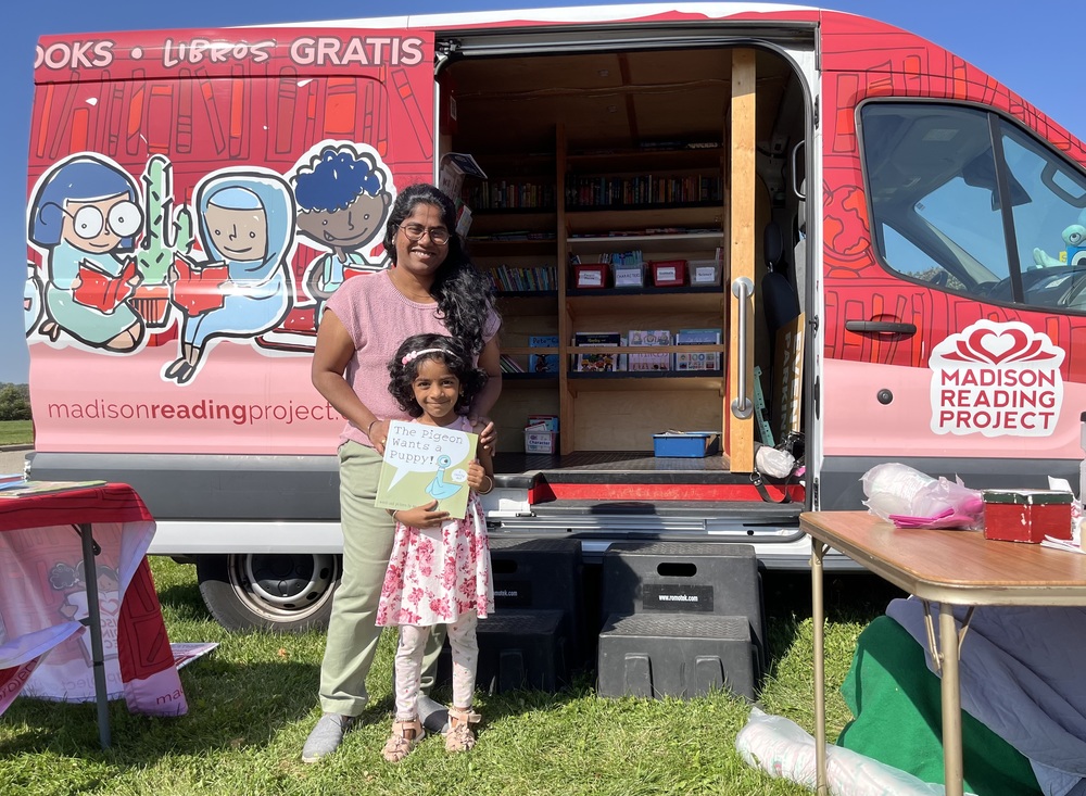 Parent and child smiling while holding books and standing in front of the Madison Reading Project book van