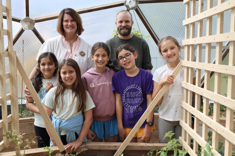 Dr. Monogue, Ren Schwab, and elementary school students smile in front of plants in a greenhouse