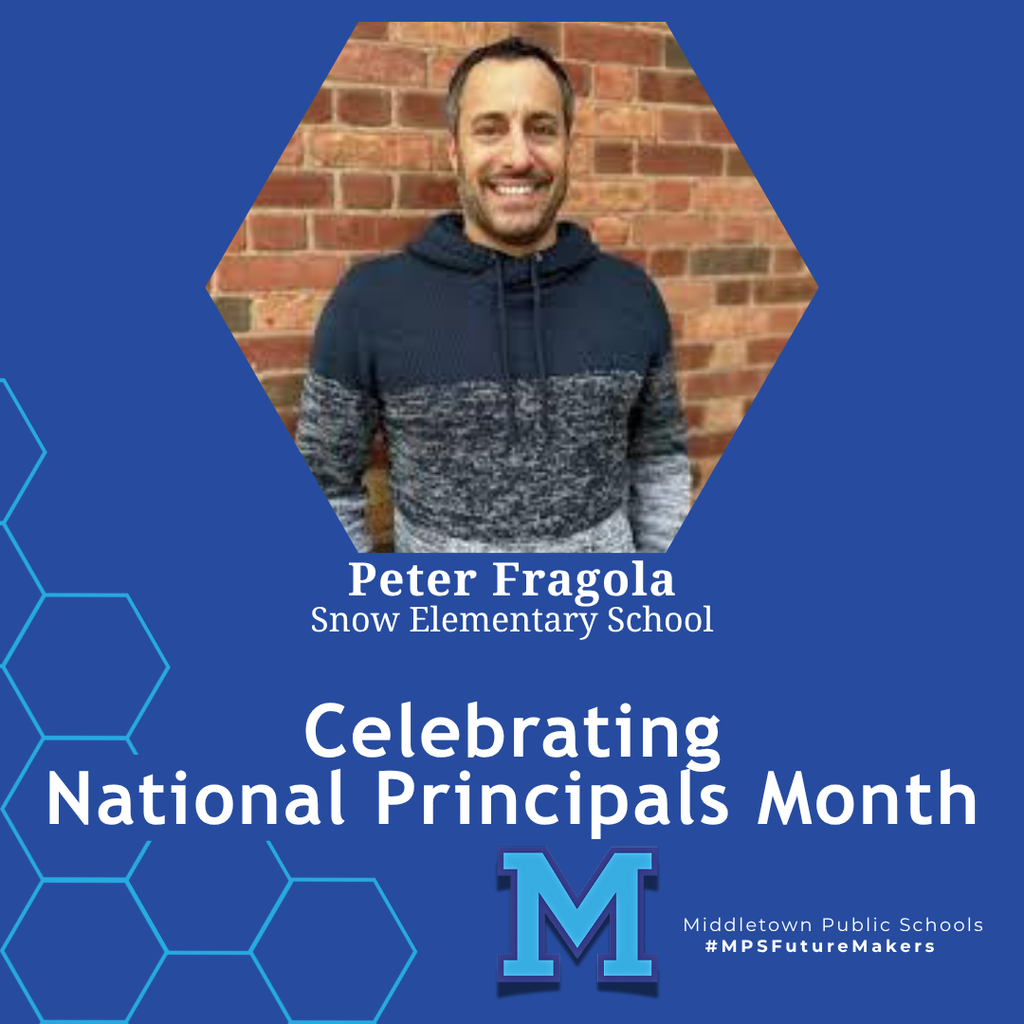 A man in a blue and grey sweatshirt, smiling and standing in front of a brick wall. The image celebrates National Principals Month and includes school branding.