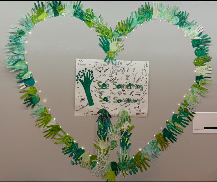 "See Something Say Something" with student signatures on a paper, surrounded by a heard made with hand cut outs and lights in a heart shape.