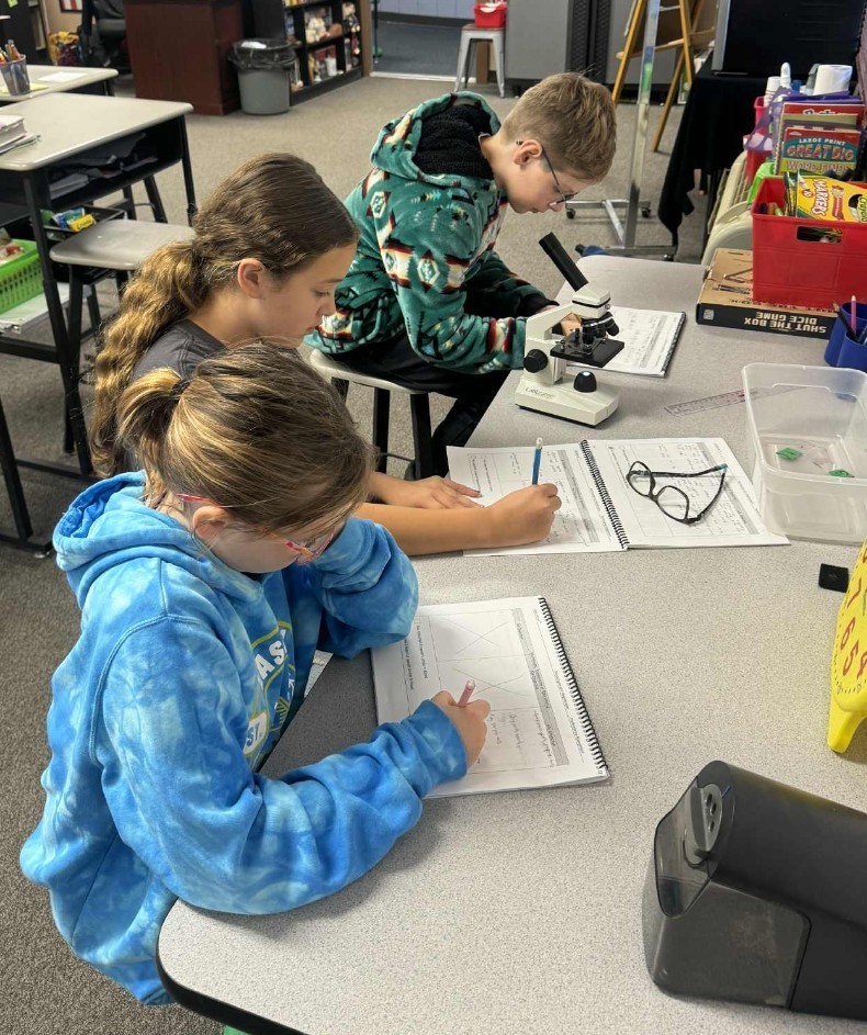3 kids looking at a microscope and writing what they see.