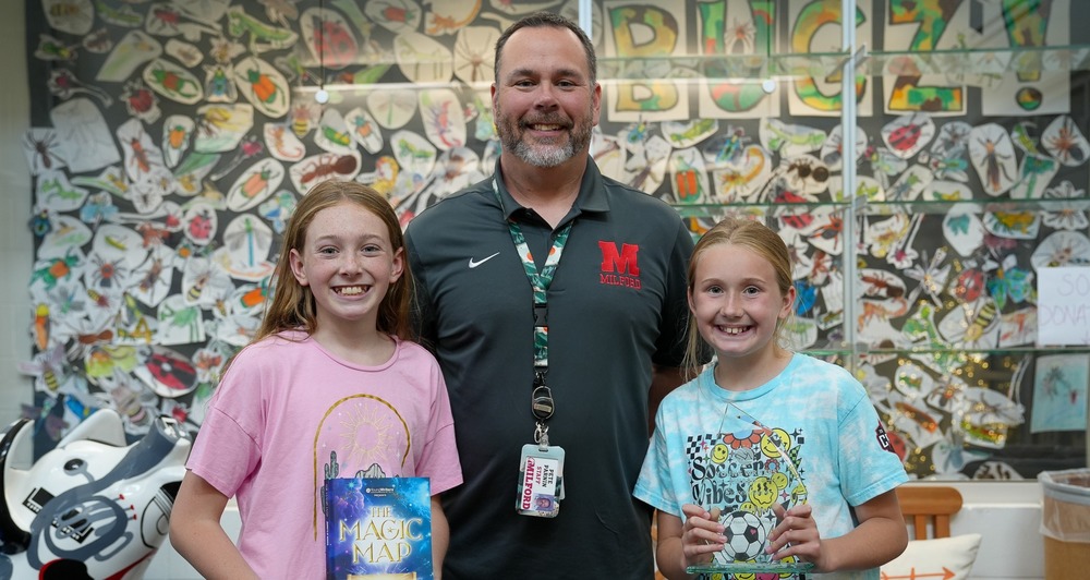 A teacher stands smiling between two elementary-aged students in a school hallway or classroom display area. The student on the left holds a book titled The Magic Map: Journeys of Wonder, while the student on the right holds a clear award plaque. All three face the camera and smile. Behind them is a colorful wall filled with student artwork featuring insects and drawings, along with the word “BUGZ” displayed across the top.