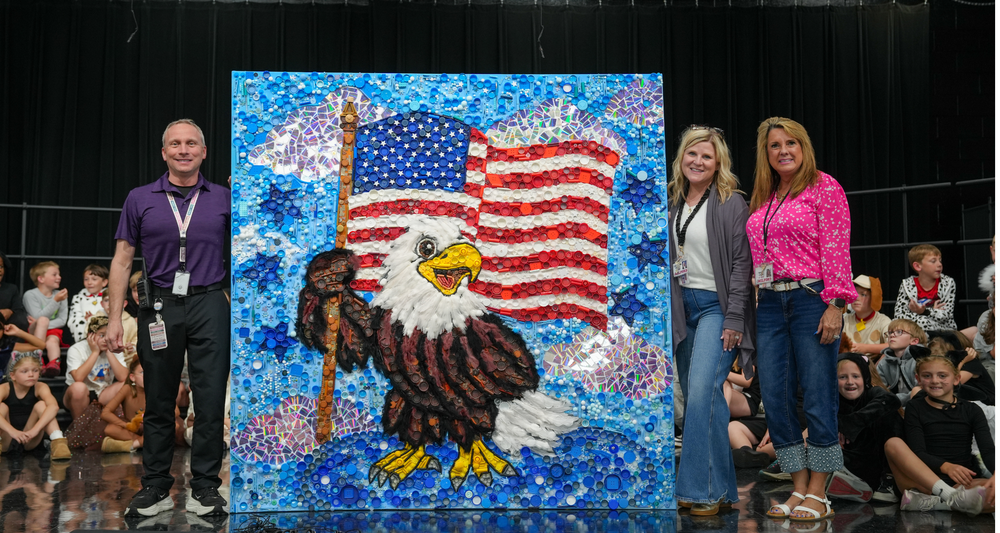 Three adults stand beside a large collaborative student mural featuring a bald eagle in front of an American flag. The artwork is made from colorful found objects such as bottle caps, beads, and tiles, creating a textured mosaic effect. Students sit in the background on a stage, watching the presentation.