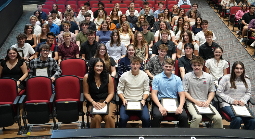 Group photo of Milford High School Class of 2026 Summa Cum Laude students seated in rows inside an auditorium. Students are dressed in semi-formal attire and smiling toward the camera. Several students in the front row hold framed academic awards while sitting in red theater seats, with additional students and a few adults visible in the background.