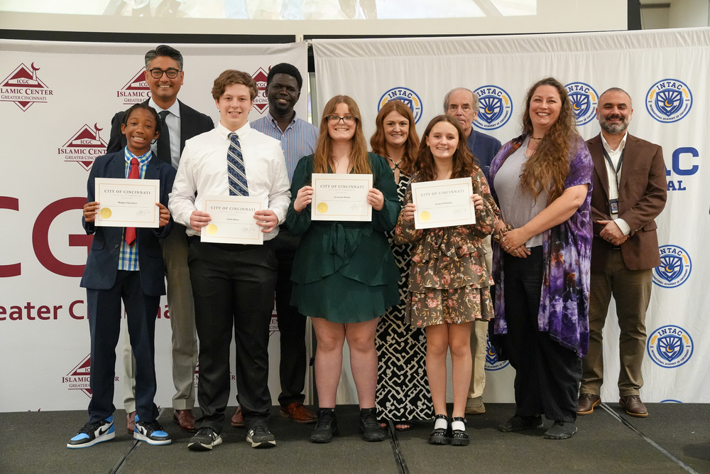 A group of Milford middle school robotics students stand on a stage holding City of Cincinnati certificates, joined by coaches and local leaders, during a recognition ceremony. The students are dressed in formal attire and smiling, with event banners displayed behind them.