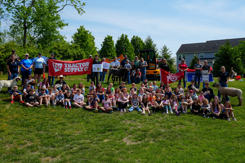 A large group of students, staff, and community members pose together on a grassy hill with farm animals and equipment during an agricultural event.