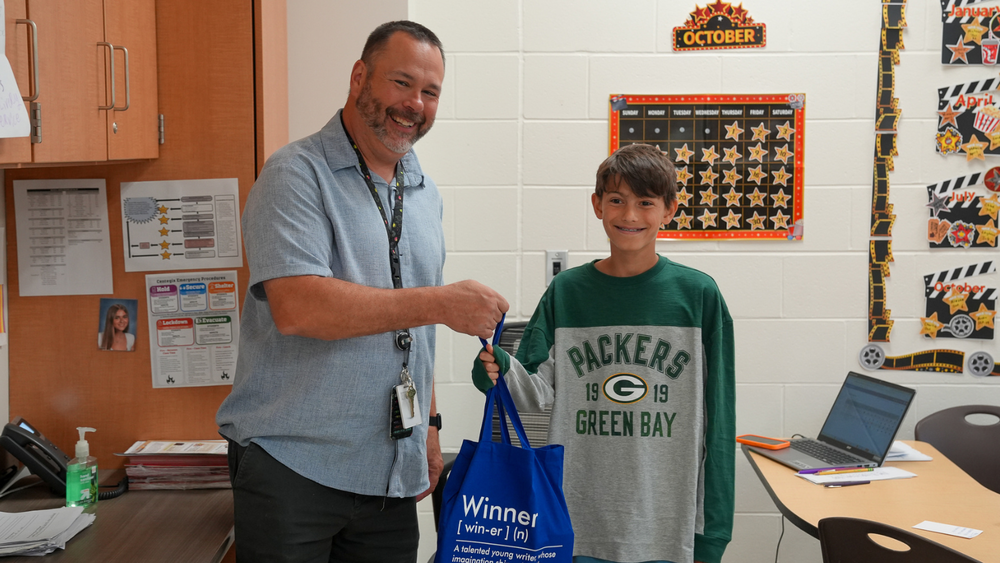 A man wearing a blue short sleeve shirt handing a blue tote bag that says "winner" to a young student wearing a greenbay packers shirt