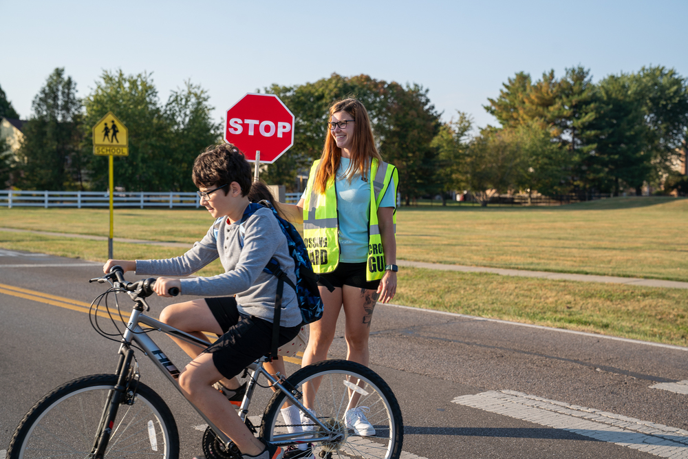 a woman with bright green crossing guard vest holding a stop sign assisting a boy riding a bike cross the street
