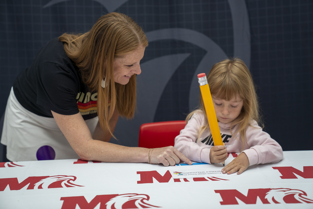 Red-Haired woman smiling as girl with big pencil writes